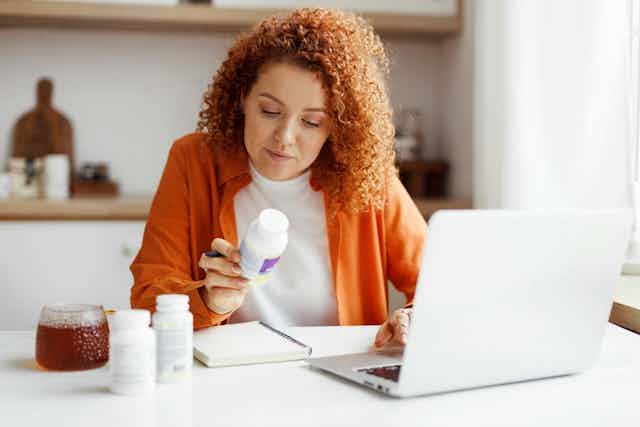 Woman looking at pill bottle while at desk with laptop open