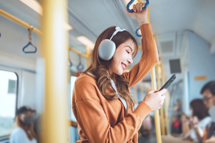 A happy young woman standing on a bus or train, wearing large headphones and looking at her mobile phone