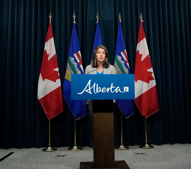 A woman stands behind a lectern that says Alberta; a row of flags are behind her.