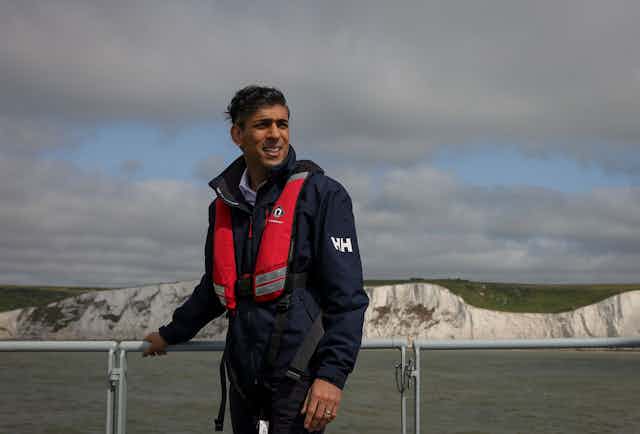 Wearing a red life vest, Rishi Sunak stands on the deck of a boat in front of the white cliffs of Dover