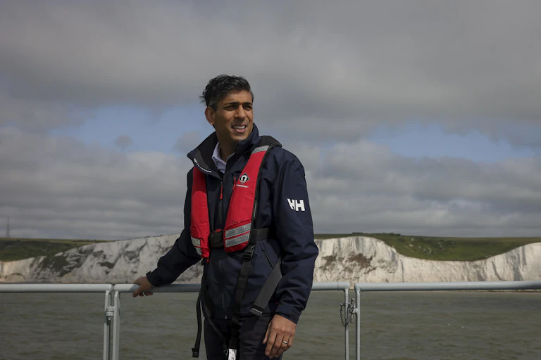 Wearing a red life vest, Rishi Sunak stands on the deck of a boat in front of the white cliffs of Dover