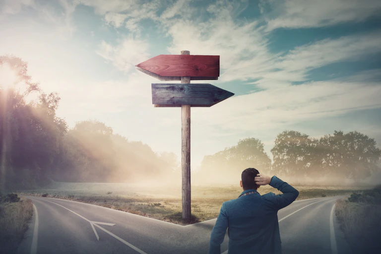 A man standing scratching his head at a junction where one path leads left and the other right. A wooden arrow point in either direction