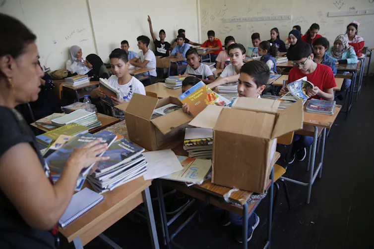 A teacher in a classroom distrbuting books from a box to students.