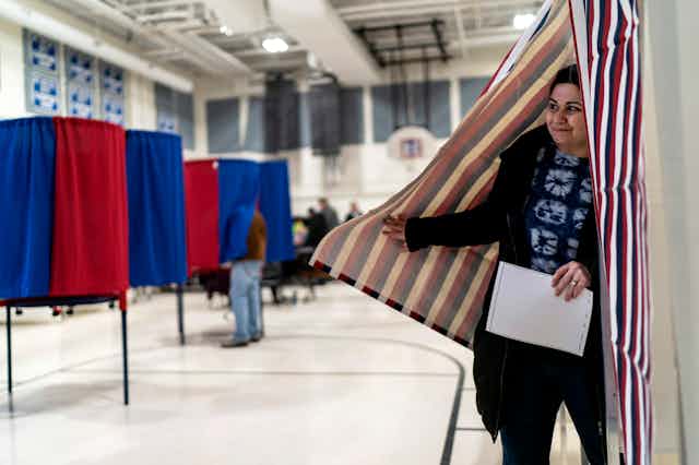 A person moves aside a curtain around a voting booth and steps out, holding a piece of paper.
