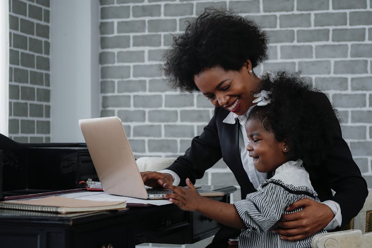 A toddler sits on the lap of a women, presumably her mother, in front of a desk. She is smiling and touching a laptop while her mother smiles down at her.
