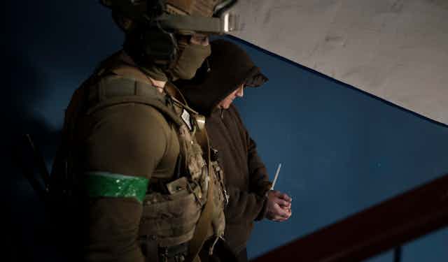 A man in a hoodie has his hands in a zip tie and is accompanied by someone in military fatigues.