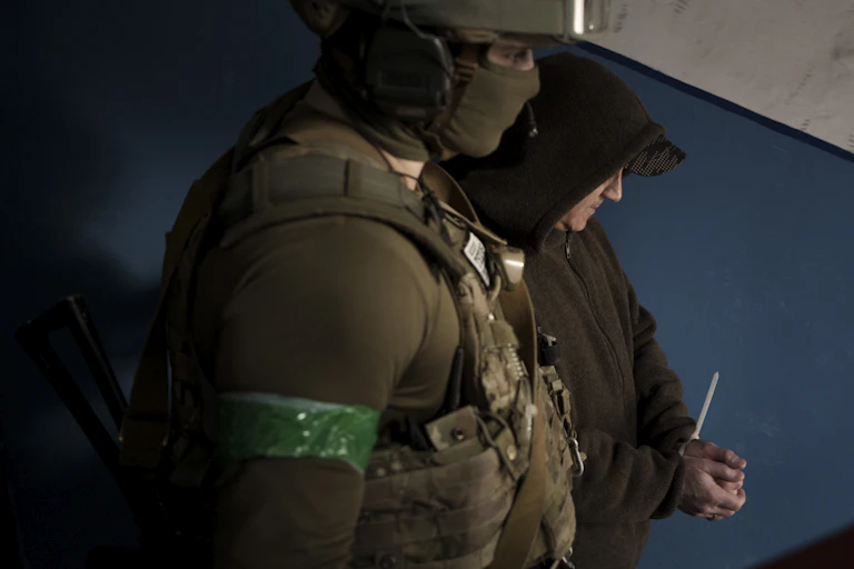 A man in a hoodie has his hands in a zip tie and is accompanied by someone in military fatigues.