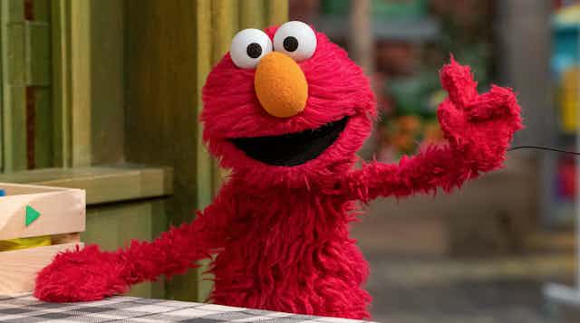 A red puppet with yellow nose and large eyes waving