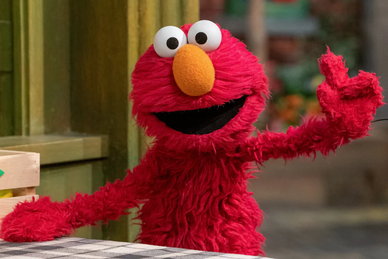 A red puppet with yellow nose and large eyes waving