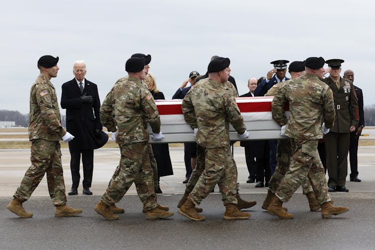 Soldiers wearing camouflage uniforms carry a casket draped in an American flag on a grey day. President Joe Biden stands nearby in a dark jacket.