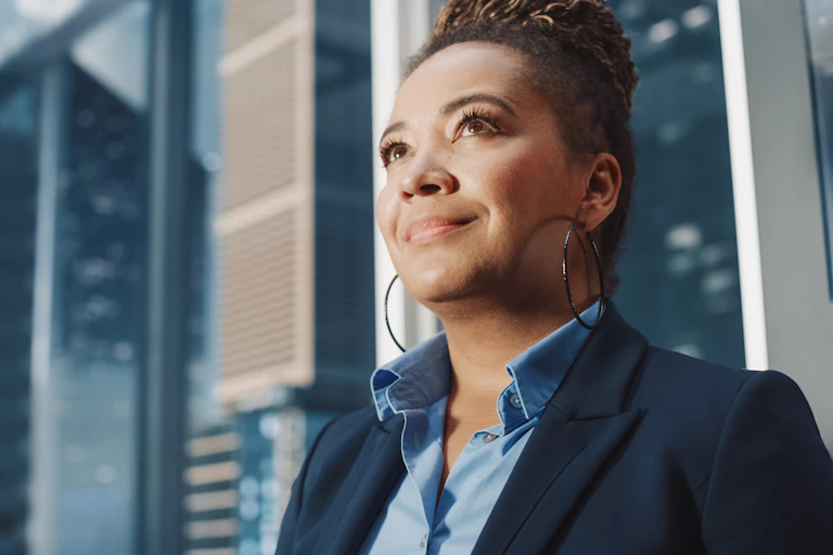 A women in business attire looks up into the distance with a small smile. Behind her, high-rise business buildings can be seen.