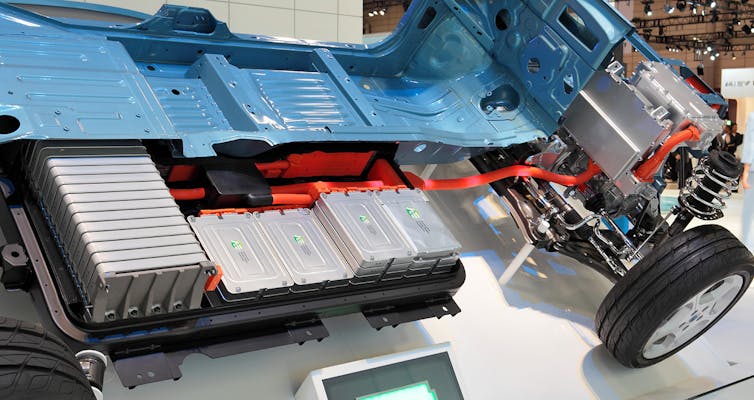 The bottom half of a Nissan Leaf electric vehicle, or a cutaway view, shows part of its battery array, a series a silver boxes, below a blue metal seat shell with red pipes running through it. 