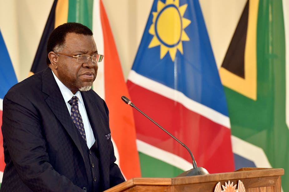 A man speak at a podium with the Namibian and South African flags in the background.