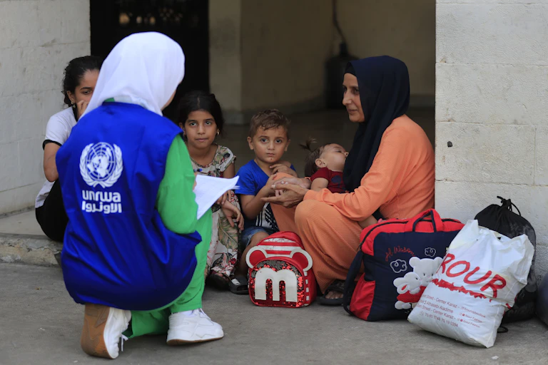 A woman wearing a blue UNRWA vest speaks to a woman with young children.