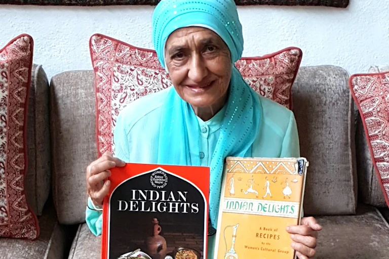 An elderly woman in a light blue headscarf and matching top proudly displays two books, different editions of Indian Delights