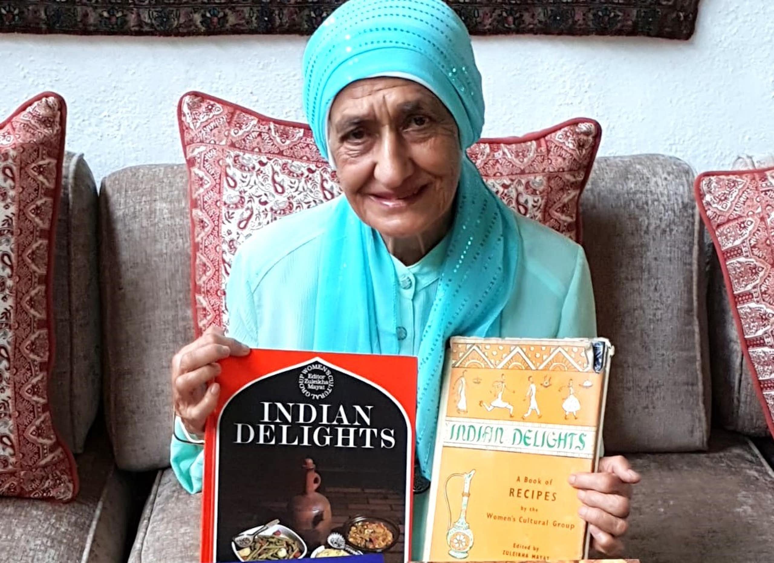 An elderly woman in a light blue headscarf and matching top proudly displays two books, different editions of Indian Delights