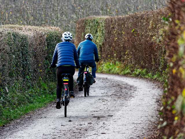 country lane, two cyclists wearring blue jackets and helmets, riding e-bikes away from camera down a hedge-lines country lane