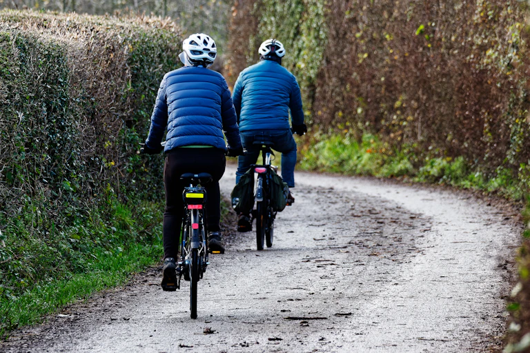 country lane, two cyclists wearring blue jackets and helmets, riding e-bikes away from camera down a hedge-lines country lane