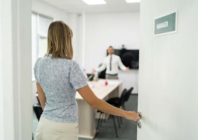 woman with hand on knob of door marked manager, man in room gestures angrily