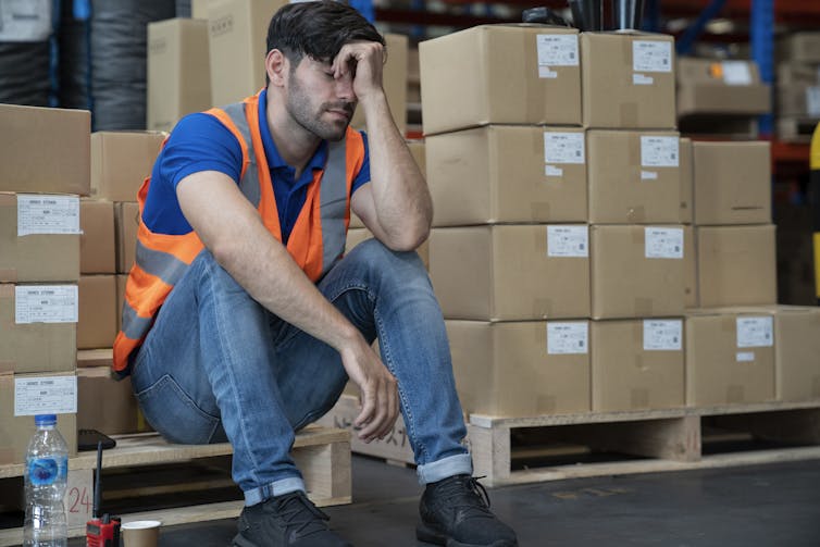 man in reflective vest sits on pallet with boxes with his head leaning on his hand