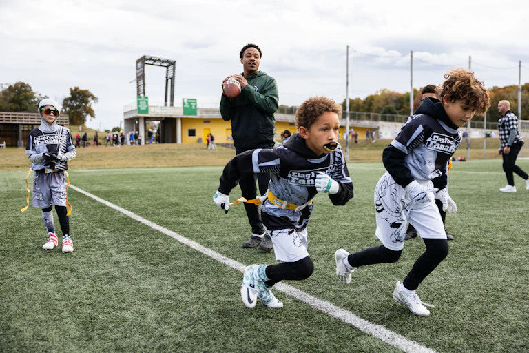 Man drops back to pass a football as two boys wearing belts with flags sprint away from him.