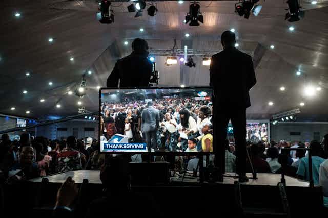 Two men stand at a giant TV screen. They're on a platform, one behind a camera, filming scenes from a gathering of many people in what appears to be a giant tent with lights. On the screen a man in a grey suit moves along the front row of people in worship.