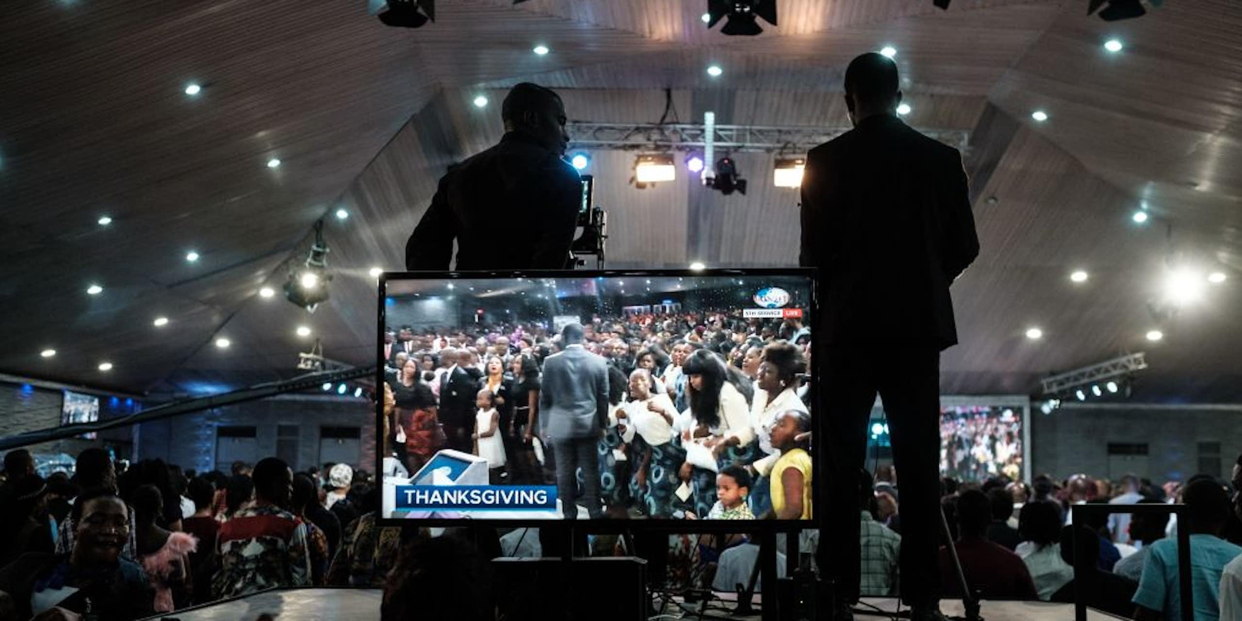 Two men stand at a giant TV screen. They're on a platform, one behind a camera, filming scenes from a gathering of many people in what appears to be a giant tent with lights. On the screen a man in a grey suit moves along the front row of people in worship.