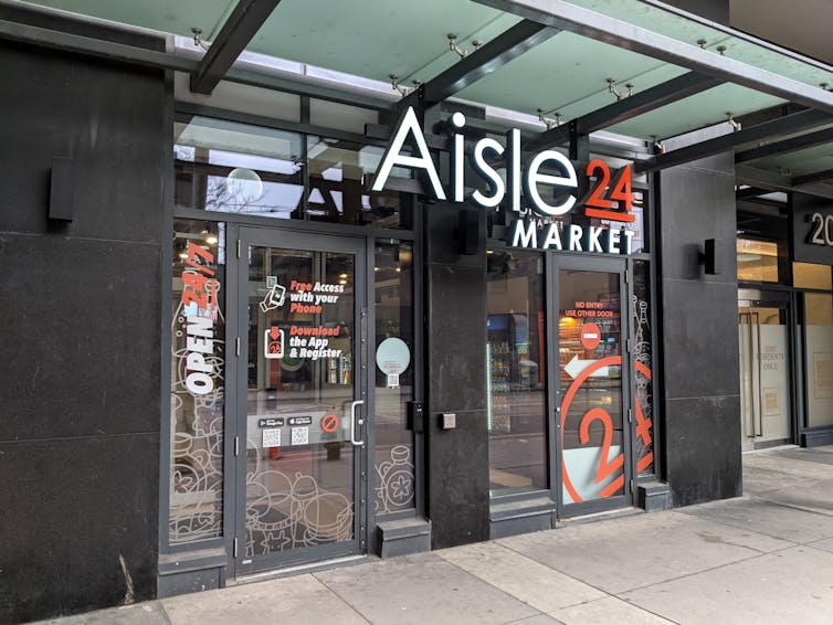 The front facade of an Aisle 24 store, with instructions reading 'Free access with your phone' and 'Download the app & register' on the front door