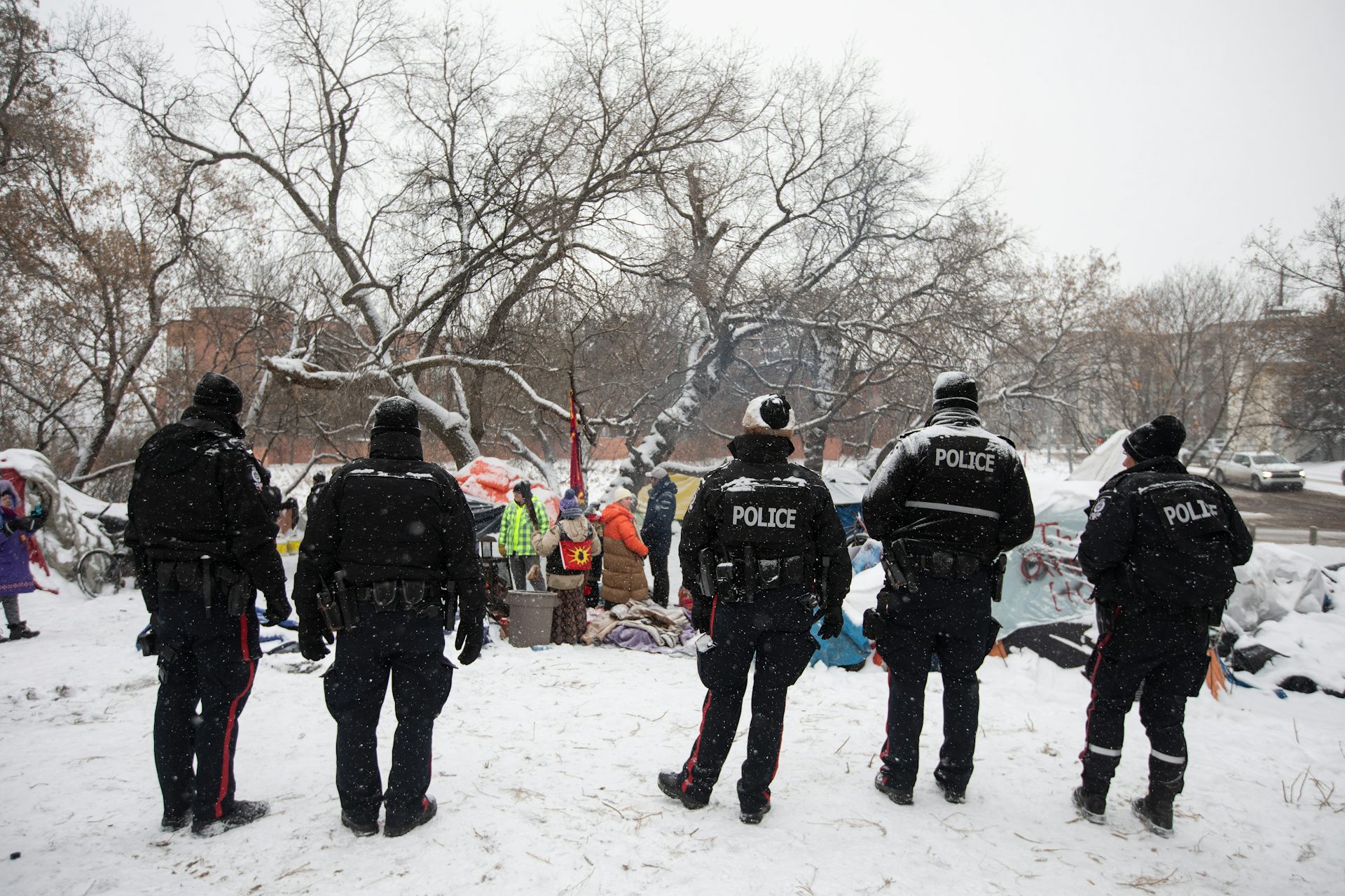 A line of police officers face a snowy homeless encampment.