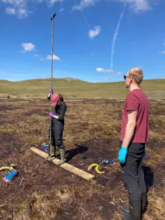 Woman and man wearing wellies and gloves, holding survey equipment on peatland bogs with green hills and blue sky in background