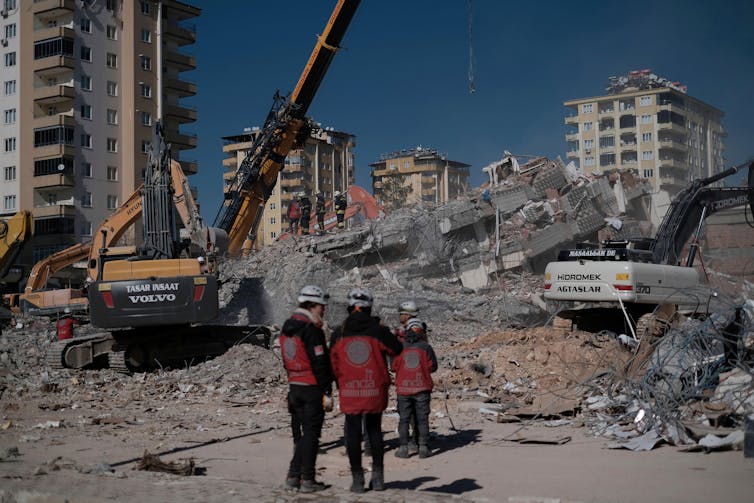Three workers in dressed in red standing in front of earthquake-damaged buildings.