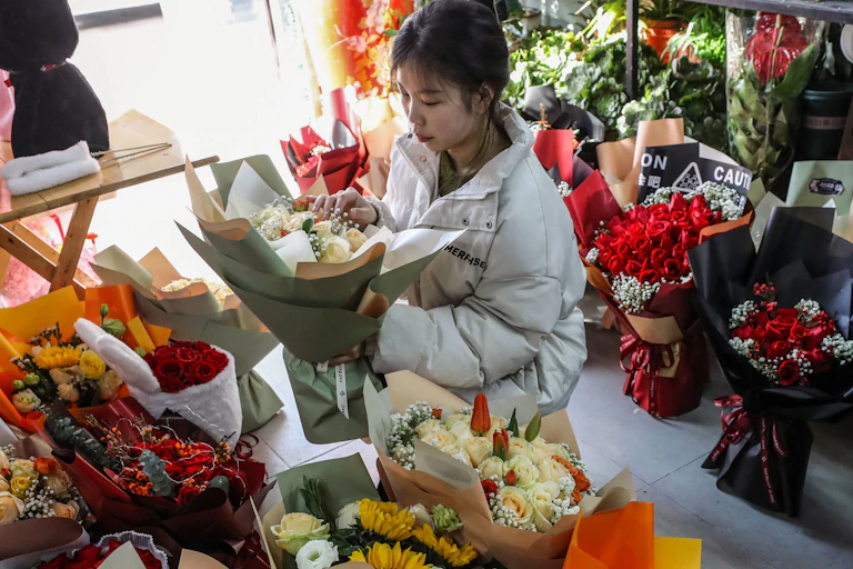 A young woman holding a bouquet of flowers while standing in a shop, surrounded by colorful bouquets ready to be sold.