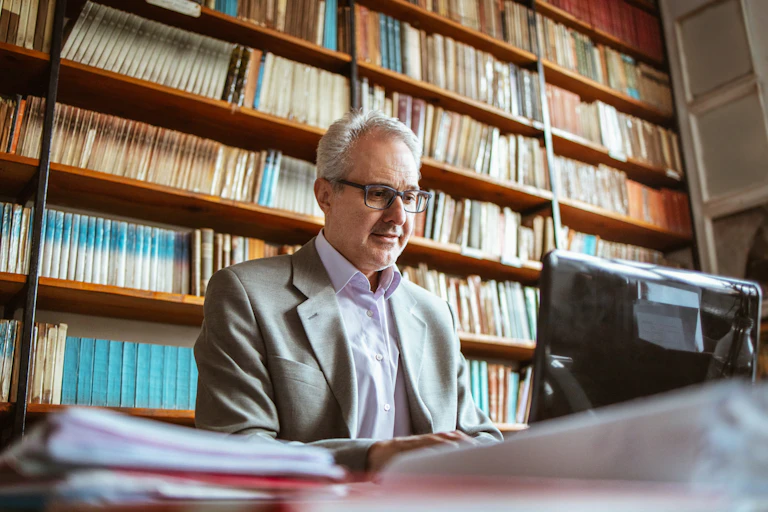 A middle aged man sits at a computer against a wall full of books.