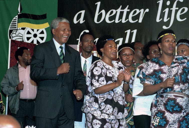 Nelson Mandela wears a dark suit and dances alongside women, in front of a sign that has the words 'a better life.'