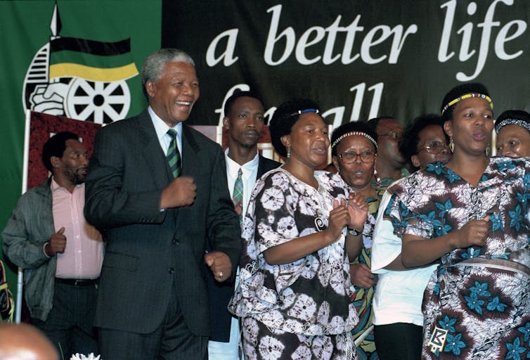 Nelson Mandela wears a dark suit and dances alongside women in front of a sign that says 