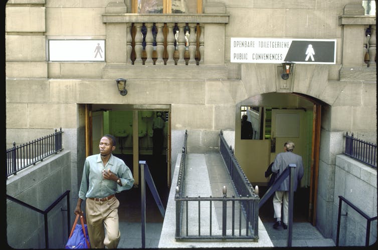 A Black man walks away from a limestone building, while a white man is seen entering on the other side. There are two signs above the entryways, one that shows a black man and the other shows a white man.