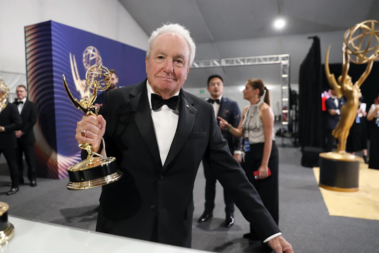 White-haired man in a tuxedo holds an Emmy award