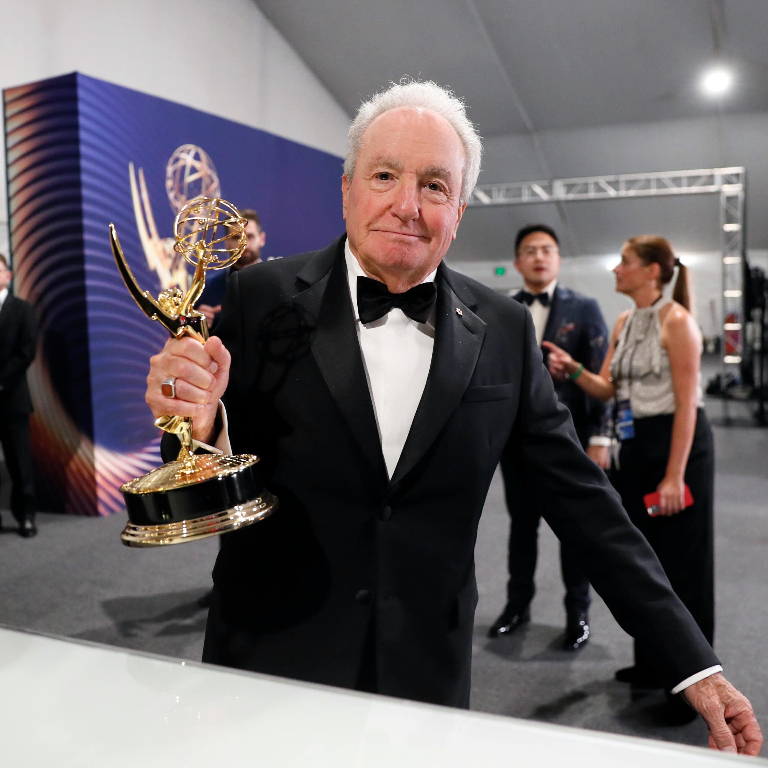 White-haired man in a tuxedo holds an Emmy award