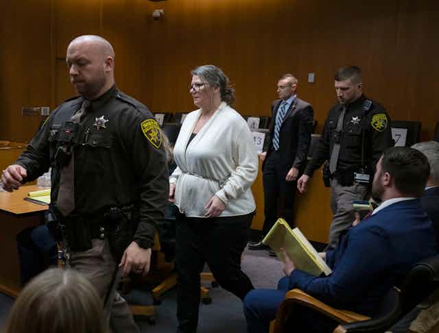 A white woman has her wrists bound in a chain as she walks between two law enforcement officers.