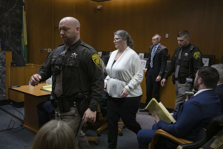 A white woman has her wrists bound in a chain as she walks between two law enforcement officers.