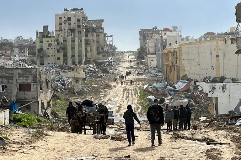 Silhouettes of people are seen at the top of a hill. Around the people are destroyed, crumbled buildings, and a dirt road.