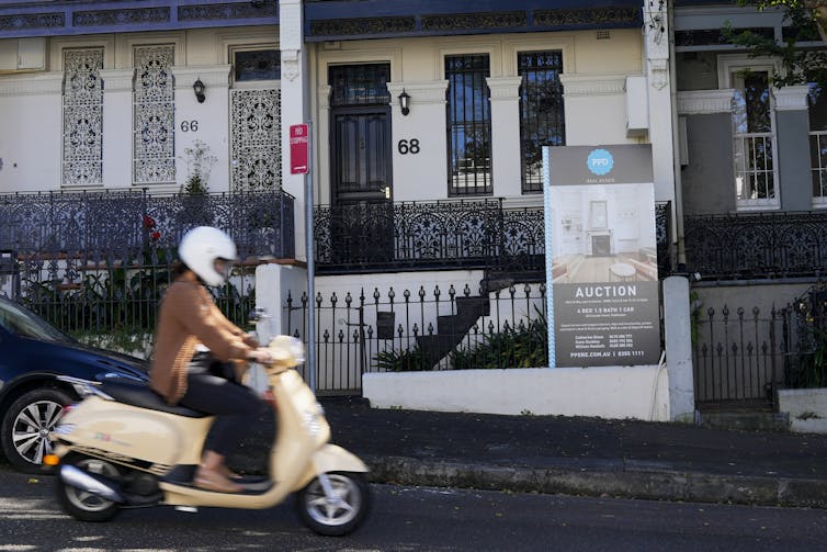 A moped driver outside a house with an auction sign in Sydney.