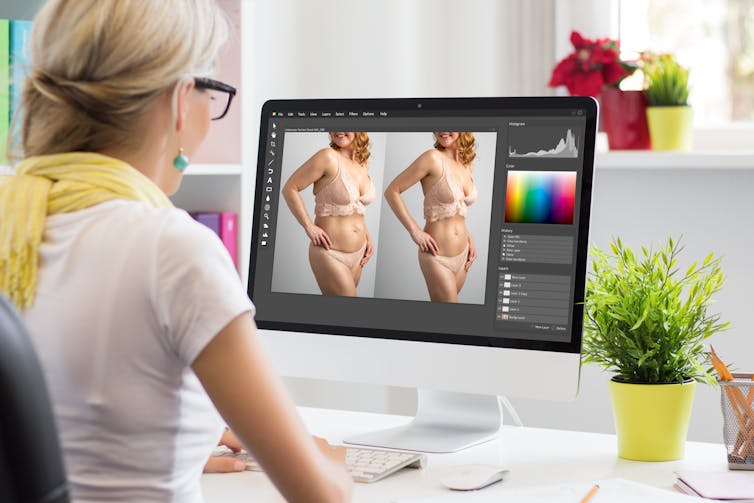 A woman sits on a computer using photo editing software to alter a photo of a woman in underwear