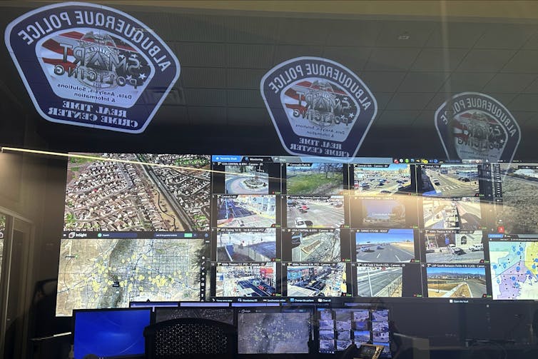 A wall of monitors shows aerial and street views of a city