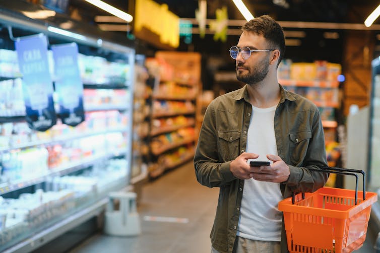 A young man stands in a supermarket holding a phone.