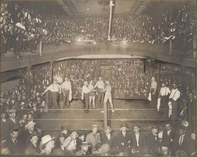 A historic scene of men in the middle of a room preparing for a boxing match surrounded by a large crowd