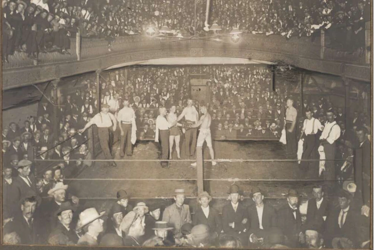 A historic scene of men in the middle of a room preparing for a boxing match surrounded by a large crowd