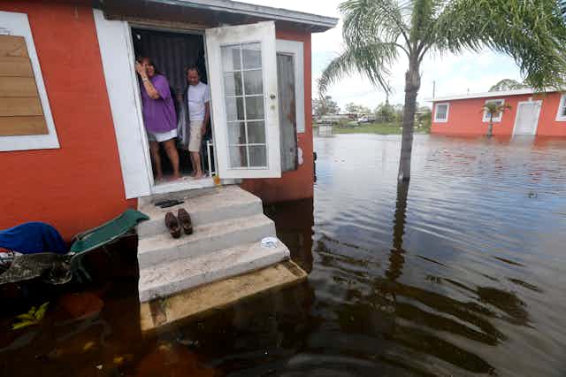 A couple stand in the doorway of their home with water surrounding it, including up the front step.