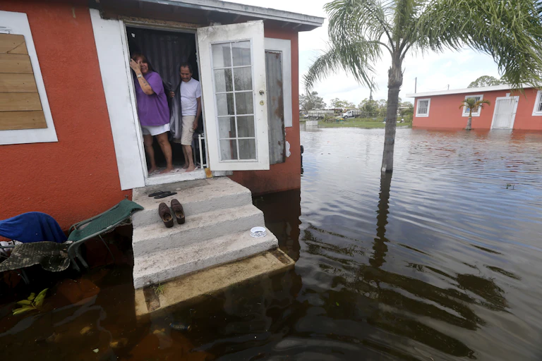 A couple stand in the doorway of their home with water surrounding it, including up the front step.