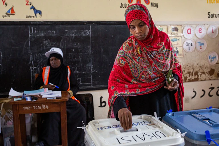 A woman wearing a headscarf places her ballot into a box in a school classroom.